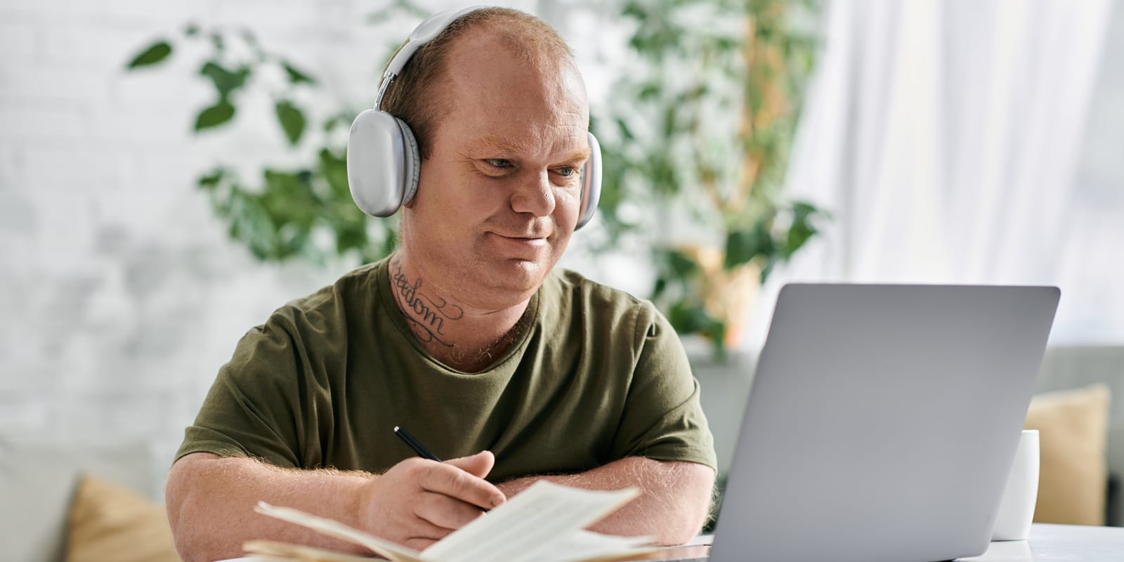 A man wearing over-ear headphones sits at a laptop with an open notebook and a pen in hand, in a bright room.