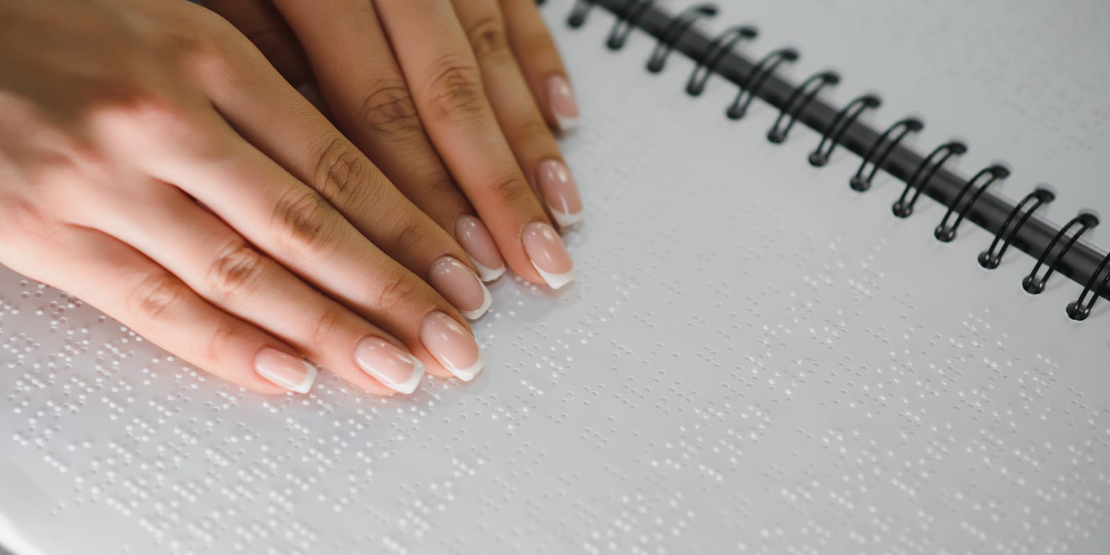 Close-up of two hands reading a page of braille in a spiral-bound book.